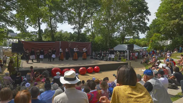 La photo montre un spectacle en plein air devant une foule assise. Une scène rectangulaire est installée sous de grands arbres. Sur scène, plusieurs personnes portent des costumes traditionnels (jupes/tabliers, gilets, coiffes) et sont alignées devant un fond de scène brun. À l’avant de la scène, plusieurs grosses sphères rouges sont posées au sol. Le public est dense, assis sur des marches ou une pente; des chapeaux sont visibles au premier plan. La lumière est ensoleillée.