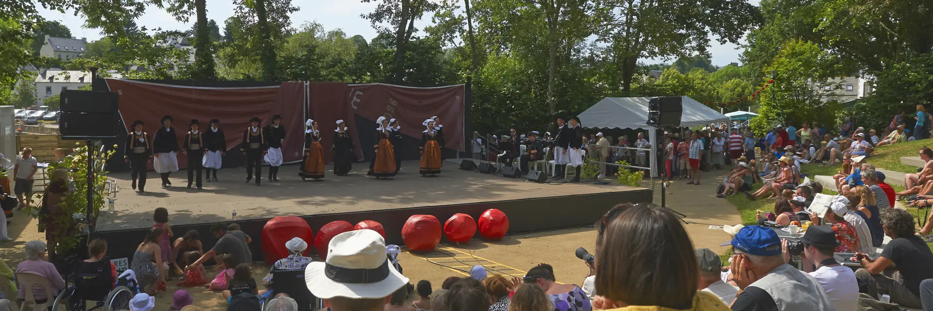 La photo montre un spectacle en plein air devant une foule assise. Une scène rectangulaire est installée sous de grands arbres. Sur scène, plusieurs personnes portent des costumes traditionnels (jupes/tabliers, gilets, coiffes) et sont alignées devant un fond de scène brun. À l’avant de la scène, plusieurs grosses sphères rouges sont posées au sol. Le public est dense, assis sur des marches ou une pente; des chapeaux sont visibles au premier plan. La lumière est ensoleillée.