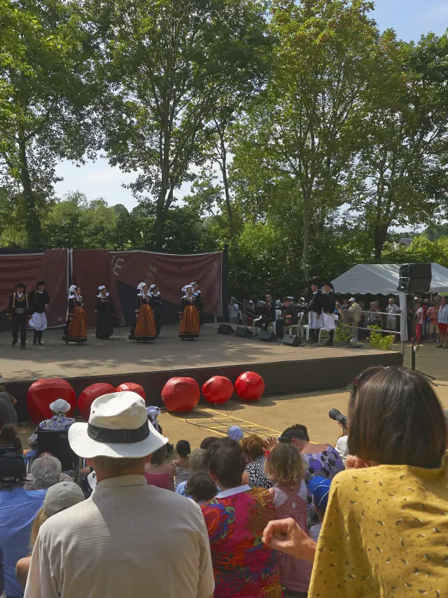 La photo montre un spectacle en plein air devant une foule assise. Une scène rectangulaire est installée sous de grands arbres. Sur scène, plusieurs personnes portent des costumes traditionnels (jupes/tabliers, gilets, coiffes) et sont alignées devant un fond de scène brun. À l’avant de la scène, plusieurs grosses sphères rouges sont posées au sol. Le public est dense, assis sur des marches ou une pente; des chapeaux sont visibles au premier plan. La lumière est ensoleillée.