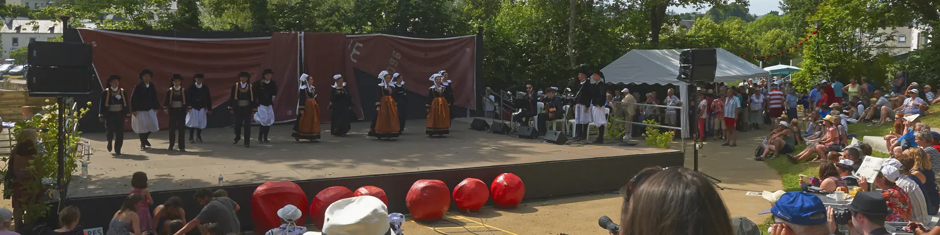 La photo montre un spectacle en plein air devant une foule assise. Une scène rectangulaire est installée sous de grands arbres. Sur scène, plusieurs personnes portent des costumes traditionnels (jupes/tabliers, gilets, coiffes) et sont alignées devant un fond de scène brun. À l’avant de la scène, plusieurs grosses sphères rouges sont posées au sol. Le public est dense, assis sur des marches ou une pente; des chapeaux sont visibles au premier plan. La lumière est ensoleillée.