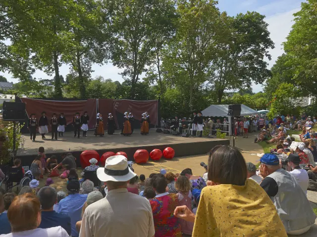 La photo montre un spectacle en plein air devant une foule assise. Une scène rectangulaire est installée sous de grands arbres. Sur scène, plusieurs personnes portent des costumes traditionnels (jupes/tabliers, gilets, coiffes) et sont alignées devant un fond de scène brun. À l’avant de la scène, plusieurs grosses sphères rouges sont posées au sol. Le public est dense, assis sur des marches ou une pente; des chapeaux sont visibles au premier plan. La lumière est ensoleillée.
