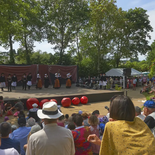 La photo montre un spectacle en plein air devant une foule assise. Une scène rectangulaire est installée sous de grands arbres. Sur scène, plusieurs personnes portent des costumes traditionnels (jupes/tabliers, gilets, coiffes) et sont alignées devant un fond de scène brun. À l’avant de la scène, plusieurs grosses sphères rouges sont posées au sol. Le public est dense, assis sur des marches ou une pente; des chapeaux sont visibles au premier plan. La lumière est ensoleillée.