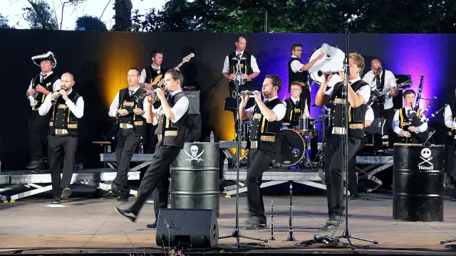 A large photo shows a group of musicians on stage (dressed in black and white), with several instruments (bagpipes, percussion, brass), microphones on stands in the foreground and stage lighting (purple, yellow/orange). Black barrels used as scenery bear white logos (including a skull) and another barrel bears the words Hénaff.
