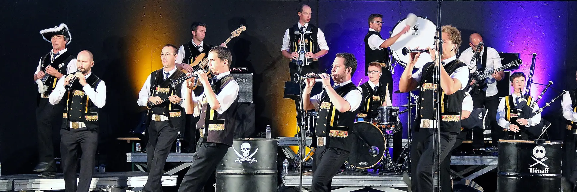 Une grande photo montre un groupe de musiciens sur scène (tenues noires et blanches), avec plusieurs instruments (cornemuses, percussions, cuivres), des micros sur pieds au premier plan et des éclairages de scène (violet, jaune/orange). Des fûts noirs servant de décor portent des logos blancs (dont un crâne) et un autre fût affiche “Hénaff”.