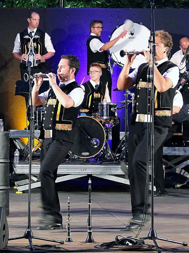 Une grande photo montre un groupe de musiciens sur scène (tenues noires et blanches), avec plusieurs instruments (cornemuses, percussions, cuivres), des micros sur pieds au premier plan et des éclairages de scène (violet, jaune/orange). Des fûts noirs servant de décor portent des logos blancs (dont un crâne) et un autre fût affiche “Hénaff”.