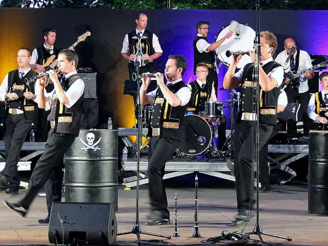 Une grande photo montre un groupe de musiciens sur scène (tenues noires et blanches), avec plusieurs instruments (cornemuses, percussions, cuivres), des micros sur pieds au premier plan et des éclairages de scène (violet, jaune/orange). Des fûts noirs servant de décor portent des logos blancs (dont un crâne) et un autre fût affiche “Hénaff”.