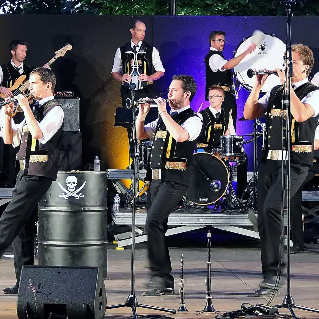 Une grande photo montre un groupe de musiciens sur scène (tenues noires et blanches), avec plusieurs instruments (cornemuses, percussions, cuivres), des micros sur pieds au premier plan et des éclairages de scène (violet, jaune/orange). Des fûts noirs servant de décor portent des logos blancs (dont un crâne) et un autre fût affiche “Hénaff”.