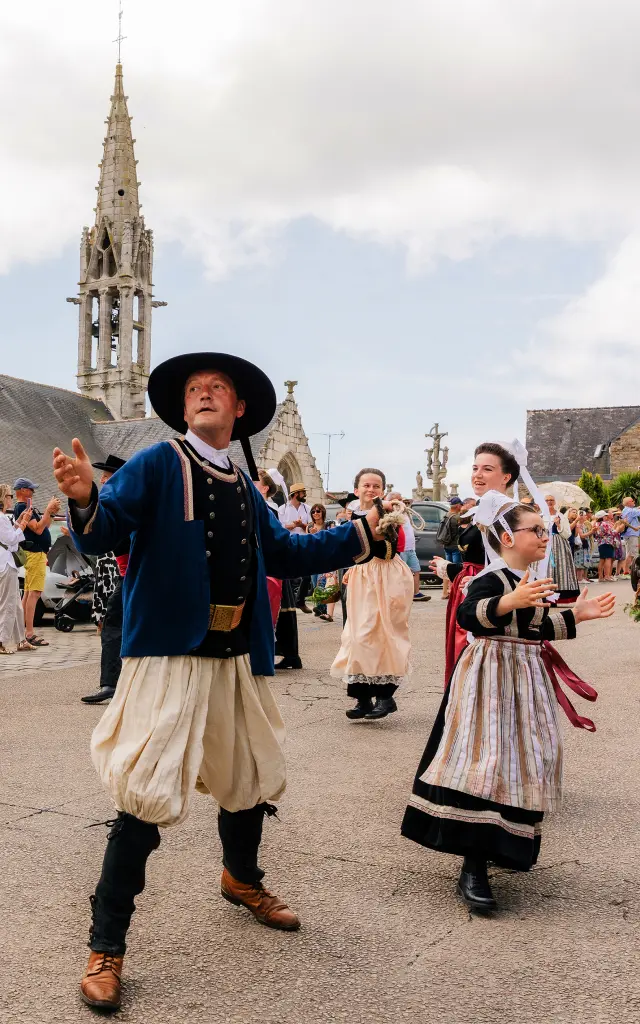 Une photo montre une scène de fête devant une église en pierre avec un clocher. Plusieurs personnes portent des costumes traditionnels et dansent sur une place; des spectateurs sont derrière des barrières. Au centre, un homme en costume traditionnel (chapeau noir, veste sombre, pantalon clair bouffant) danse, et à droite une femme en robe traditionnelle danse aussi.