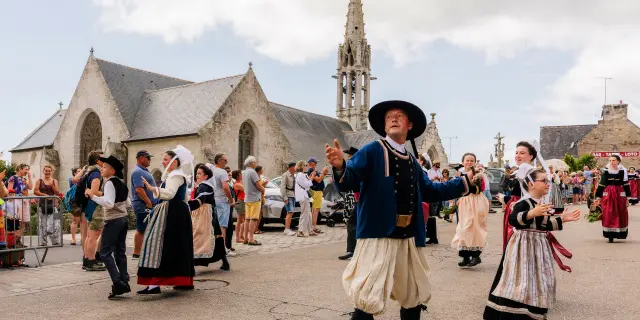 Ein Foto zeigt eine Festszene vor einer Steinkirche mit einem Glockenturm. Mehrere Personen tragen traditionelle Trachten und tanzen auf einem Platz; Zuschauer stehen hinter Absperrungen. In der Mitte tanzt ein Mann in traditioneller Tracht (schwarzer Hut, dunkle Jacke, helle, ausgebeulte Hose), rechts tanzt eine Frau in einem traditionellen Kleid.