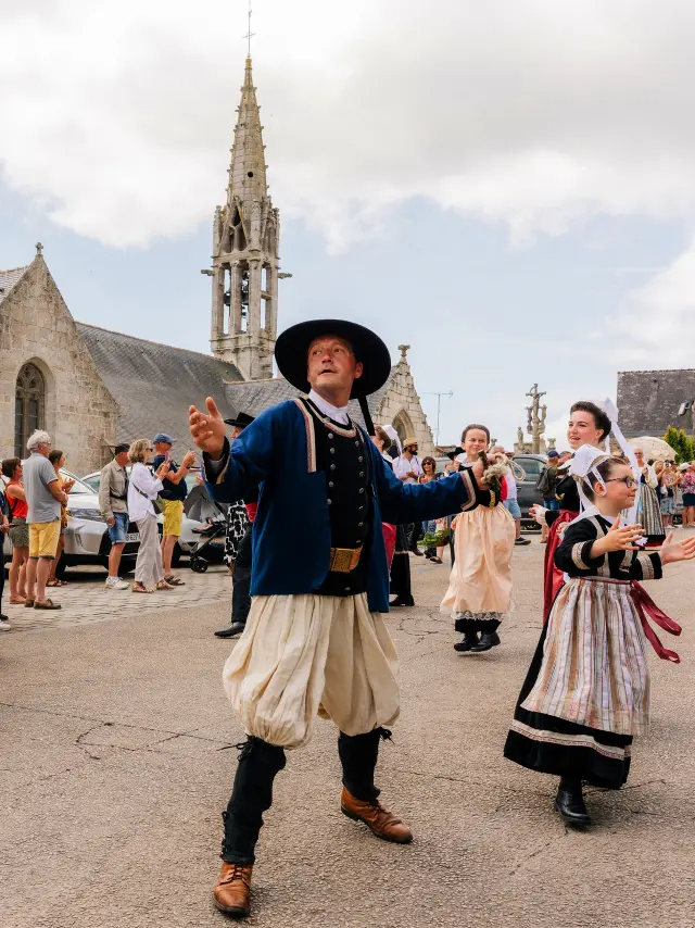 Eén foto toont een feestelijk tafereel voor een stenen kerk met klokkentoren. Verschillende mensen in traditionele klederdracht dansen op een plein, met toeschouwers achter barrières. In het midden danst een man in klederdracht (zwarte hoed, donker jasje, lichte broek) en rechts danst ook een vrouw in klederdracht.