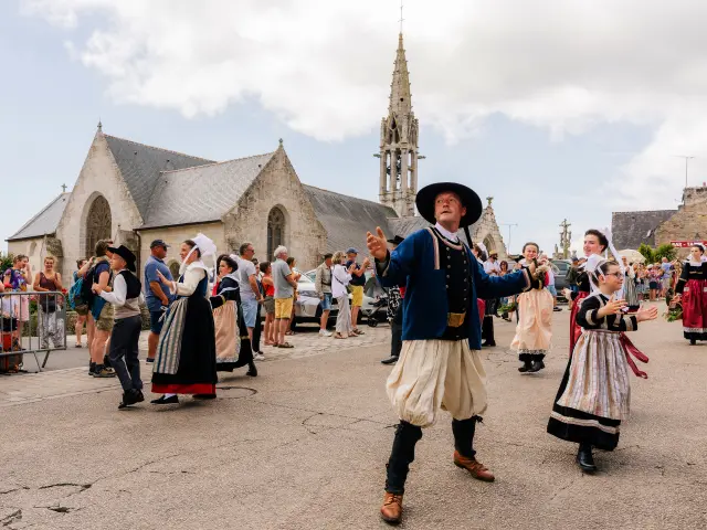 Eén foto toont een feestelijk tafereel voor een stenen kerk met klokkentoren. Verschillende mensen in traditionele klederdracht dansen op een plein, met toeschouwers achter barrières. In het midden danst een man in klederdracht (zwarte hoed, donker jasje, lichte broek) en rechts danst ook een vrouw in klederdracht.