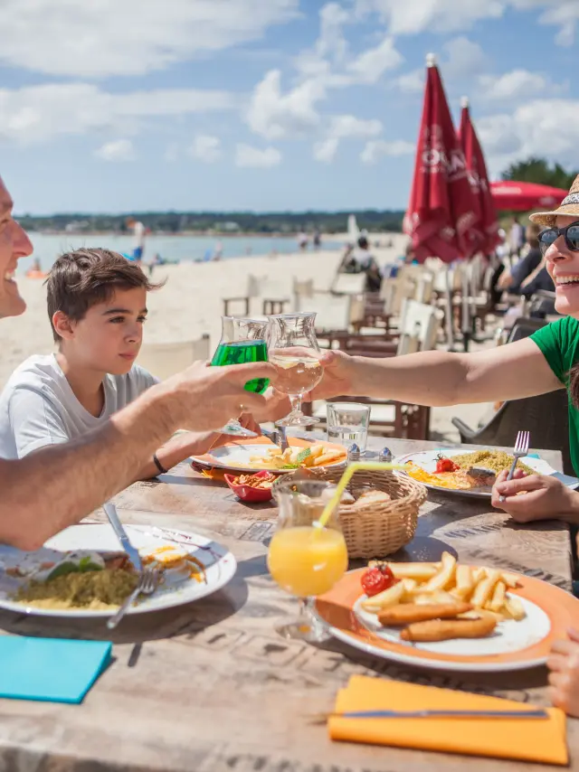 La photo montre un repas en terrasse au bord d’une plage. Quatre personnes sont assises autour d’une table avec des assiettes (frites, salade/plat), des verres et une boisson orange avec une paille. Un homme en polo bleu trinque avec une femme en haut vert portant un chapeau de paille et des lunettes de soleil. Un garçon est assis entre eux. Au premier plan à droite, un enfant porte un chapeau clair avec une fleur rouge et des lunettes vertes. En arrière-plan, on distingue la plage, la mer, des parasols rouges et des tables de restaurant.