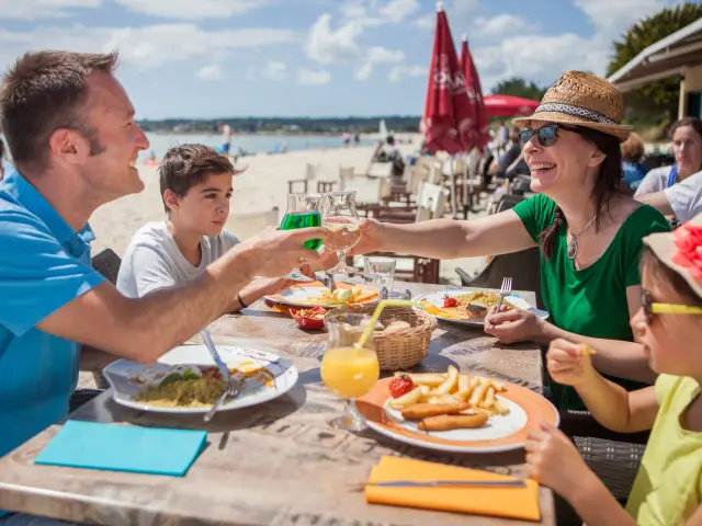 La photo montre un repas en terrasse au bord d’une plage. Quatre personnes sont assises autour d’une table avec des assiettes (frites, salade/plat), des verres et une boisson orange avec une paille. Un homme en polo bleu trinque avec une femme en haut vert portant un chapeau de paille et des lunettes de soleil. Un garçon est assis entre eux. Au premier plan à droite, un enfant porte un chapeau clair avec une fleur rouge et des lunettes vertes. En arrière-plan, on distingue la plage, la mer, des parasols rouges et des tables de restaurant.