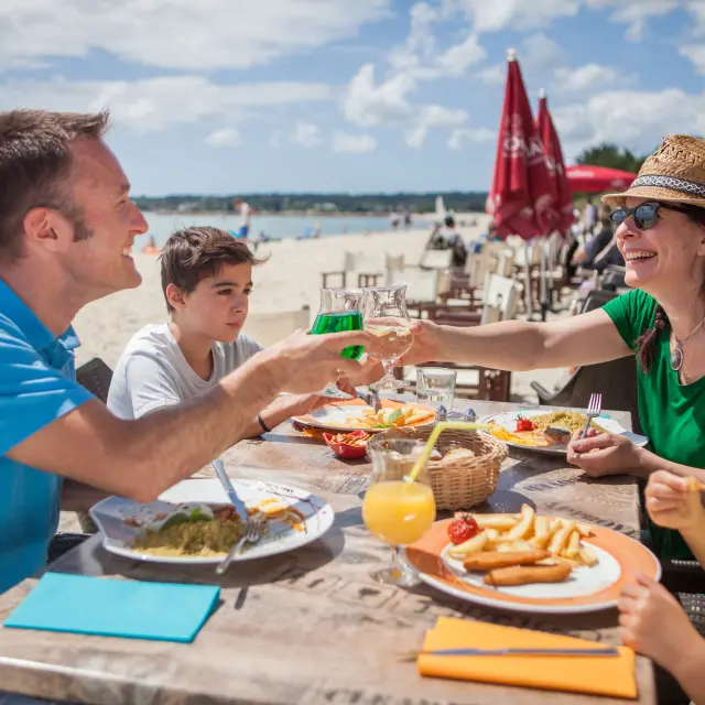 Das Foto zeigt ein Essen auf einer Terrasse an einem Strand. Vier Personen sitzen um einen Tisch mit Tellern (Pommes frites, Salat/Teller), Gläsern und einem orangefarbenen Getränk mit Strohhalm. Ein Mann in einem blauen Poloshirt stößt mit einer Frau in einem grünen Oberteil an, die einen Strohhut und eine Sonnenbrille trägt. Ein Junge sitzt zwischen ihnen. Im Vordergrund rechts trägt ein Kind einen hellen Hut mit einer roten Blume und eine grüne Brille. Im Hintergrund sind der Strand, das Meer, rote Sonnenschirme und Restauranttische zu erkennen.