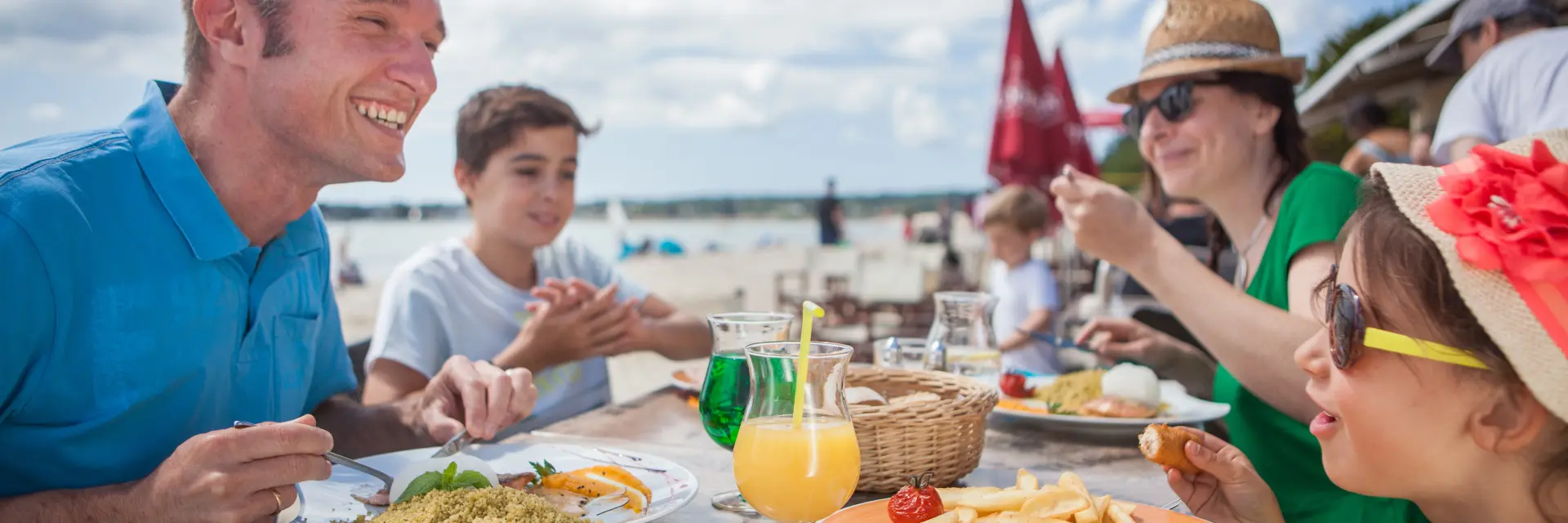 La photo montre un repas en extérieur au bord de la mer. Plusieurs personnes sont assises autour d’une table avec des assiettes (frites, salade, plat en sauce) et des boissons (un jus orange, une boisson verte). À l’avant droit, une enfant porte des lunettes de soleil et un chapeau avec une grande décoration rouge. À gauche, un homme en polo bleu sourit. En arrière-plan, la plage et le ciel avec des nuages.