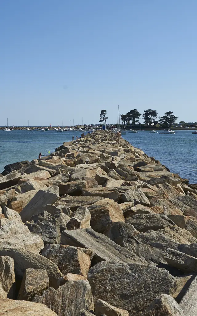 Une photo qui montre une jetée en enrochements (gros blocs de roches) qui s’avance au centre vers la mer, avec de l’eau bleue/verte de chaque côté, une plage sableuse à droite, quelques personnes près de l’eau à gauche, et un ciel bleu dégagé. La jetée mène vers une zone au loin avec quelques arbres et des constructions basses.