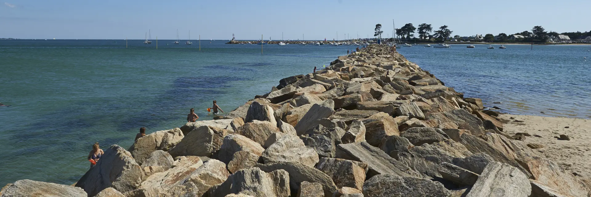 Ein Foto, das einen Pier aus Steinschüttungen (große Felsblöcke) zeigt, der in der Mitte ins Meer ragt, mit blauem/grünem Wasser auf beiden Seiten, einem Sandstrand auf der rechten Seite, einigen Menschen in Wassernähe auf der linken Seite und einem klaren blauen Himmel. Der Steg führt zu einem Gebiet in der Ferne mit einigen Bäumen und niedrigen Gebäuden.