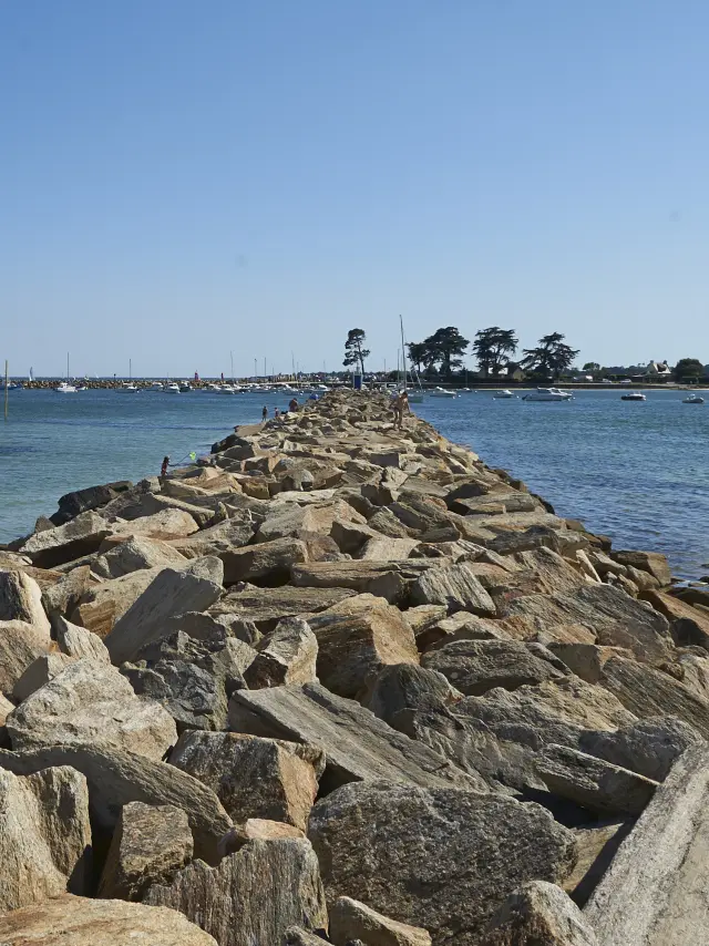 A photo showing a riprap jetty (large boulders) jutting out into the sea in the centre, with blue/green water on either side, a sandy beach on the right, a few people near the water on the left, and a clear blue sky. The jetty leads to an area in the distance with a few trees and low buildings.
