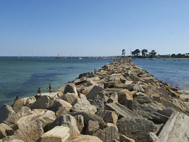 Ein Foto, das einen Pier aus Steinschüttungen (große Felsblöcke) zeigt, der in der Mitte ins Meer ragt, mit blauem/grünem Wasser auf beiden Seiten, einem Sandstrand auf der rechten Seite, einigen Menschen in Wassernähe auf der linken Seite und einem klaren blauen Himmel. Der Steg führt zu einem Gebiet in der Ferne mit einigen Bäumen und niedrigen Gebäuden.