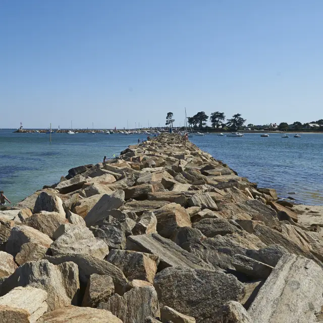Ein Foto, das einen Pier aus Steinschüttungen (große Felsblöcke) zeigt, der in der Mitte ins Meer ragt, mit blauem/grünem Wasser auf beiden Seiten, einem Sandstrand auf der rechten Seite, einigen Menschen in Wassernähe auf der linken Seite und einem klaren blauen Himmel. Der Steg führt zu einem Gebiet in der Ferne mit einigen Bäumen und niedrigen Gebäuden.