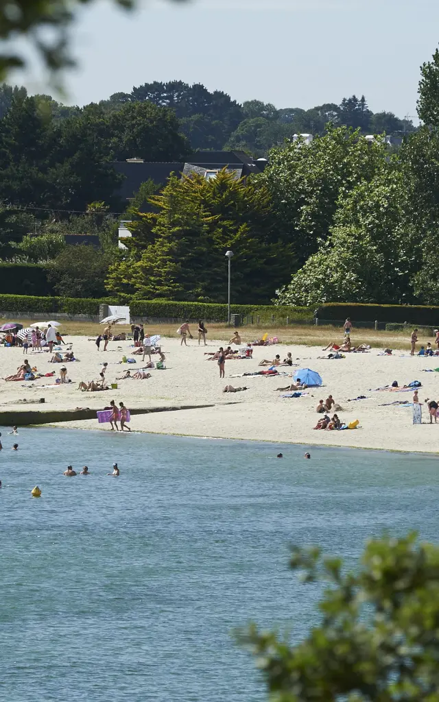Une photo de plage de sable avec beaucoup de personnes allongées ou debout, des parasols, et des baigneurs dans l’eau au premier plan; des arbres et des maisons/bâtiments en arrière-plan.