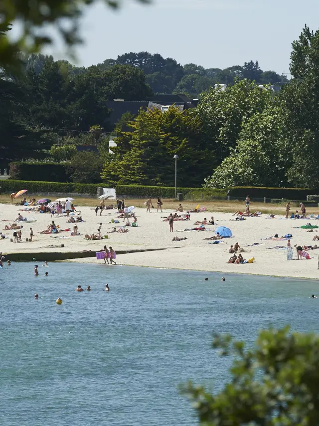 Une photo de plage de sable avec beaucoup de personnes allongées ou debout, des parasols, et des baigneurs dans l’eau au premier plan; des arbres et des maisons/bâtiments en arrière-plan.