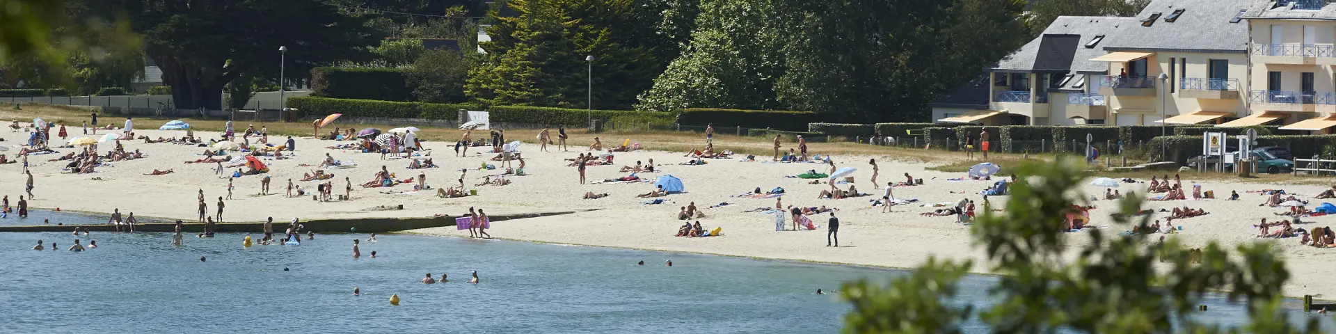 Ein Foto von einem Sandstrand mit vielen liegenden oder stehenden Menschen, Sonnenschirmen und Badenden im Wasser im Vordergrund; Bäume und Häuser/Gebäude im Hintergrund.