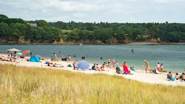 Ein Foto von einem Sandstrand mit vielen Menschen (Handtücher, Sonnenschirme) und anderen im Wasser; im Vordergrund hohes Gras; gegenüber ein bewaldeter Streifen an der Küste unter einem bewölkten Himmel.