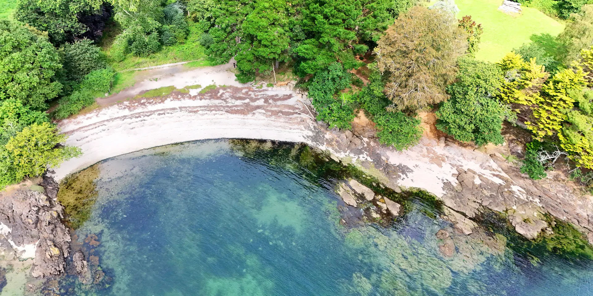 Ein Luftbild einer kleinen Bucht, das einen Streifen hellen Sand, Felsen und klares blaugrünes Wasser zeigt. Darüber befindet sich ein dicht bewaldeter Bereich mit grünen Bäumen und etwas dunklerem Laub; rechts ist ein heller Rasen mit einer sichtbaren weißen Bank.