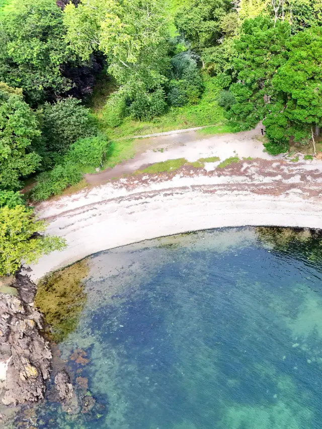 Ein Luftbild einer kleinen Bucht, das einen Streifen hellen Sand, Felsen und klares blaugrünes Wasser zeigt. Darüber befindet sich ein dicht bewaldeter Bereich mit grünen Bäumen und etwas dunklerem Laub; rechts ist ein heller Rasen mit einer sichtbaren weißen Bank.