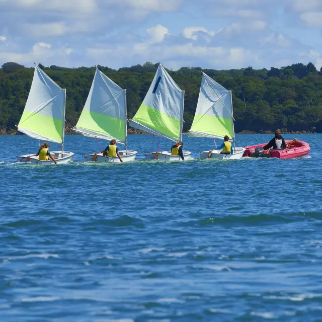 De foto toont een blauw water met kleine golven; in de verte een strook groen bos onder een gedeeltelijk bewolkte blauwe lucht. Op het water varen vier kleine rubberbootjes (witte zeilen met een lichtgroene streep) in een lijn vooruit; elk bootje lijkt te worden bezet door een persoon met een geel vest aan. Rechts van de rubberbootjes volgt een klein roze bootje met twee mensen aan boord.