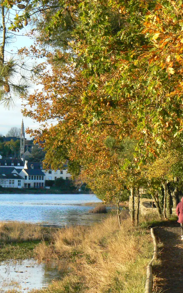 Photo d'un chemin de promenade en bord d’eau, avec des arbres aux feuilles d’automne (jaune/orange/vert) au-dessus du chemin, un muret à droite, et deux personnes marchant sur le chemin. À gauche, l’eau et, au fond, des maisons blanches d’un village.