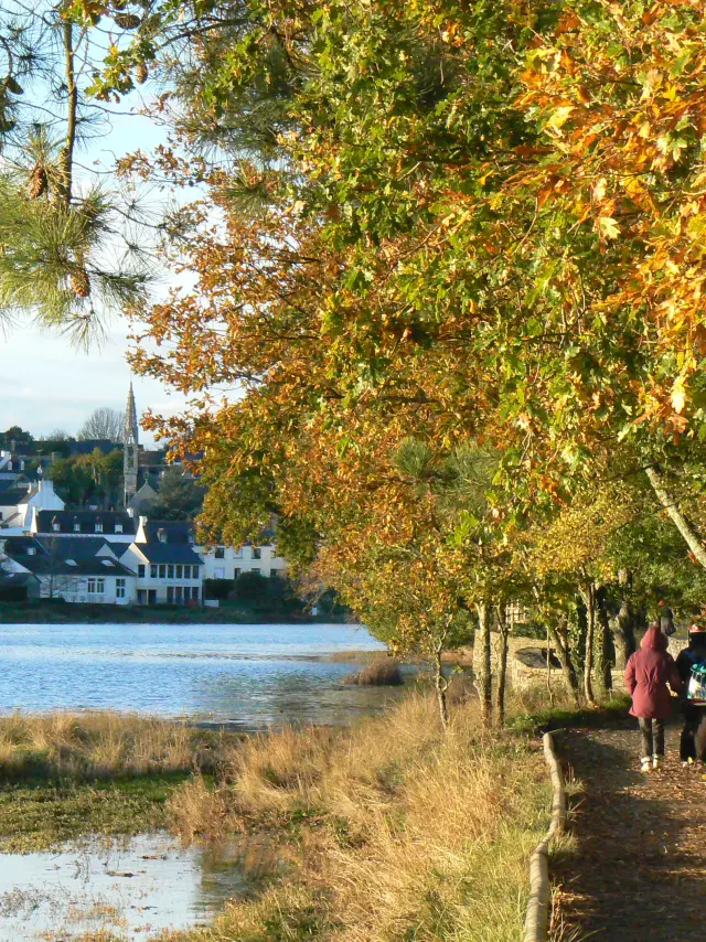 Photo d'un chemin de promenade en bord d’eau, avec des arbres aux feuilles d’automne (jaune/orange/vert) au-dessus du chemin, un muret à droite, et deux personnes marchant sur le chemin. À gauche, l’eau et, au fond, des maisons blanches d’un village.