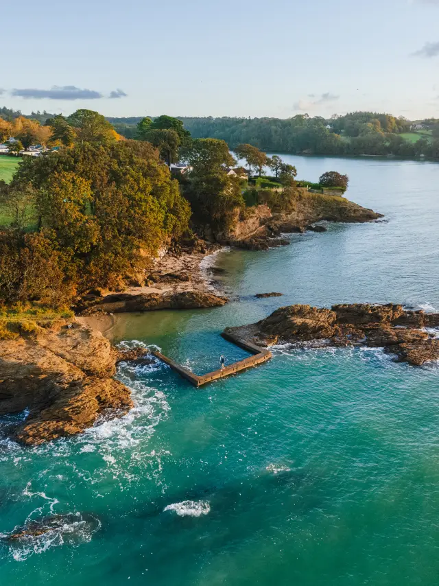 Photo de côte avec une anse avec eau turquoise, rochers, petite piscine en angle, et une bande de terre verdoyante avec des arbres aux couleurs d’automne; horizon boisé et ciel clair avec quelques nuages.