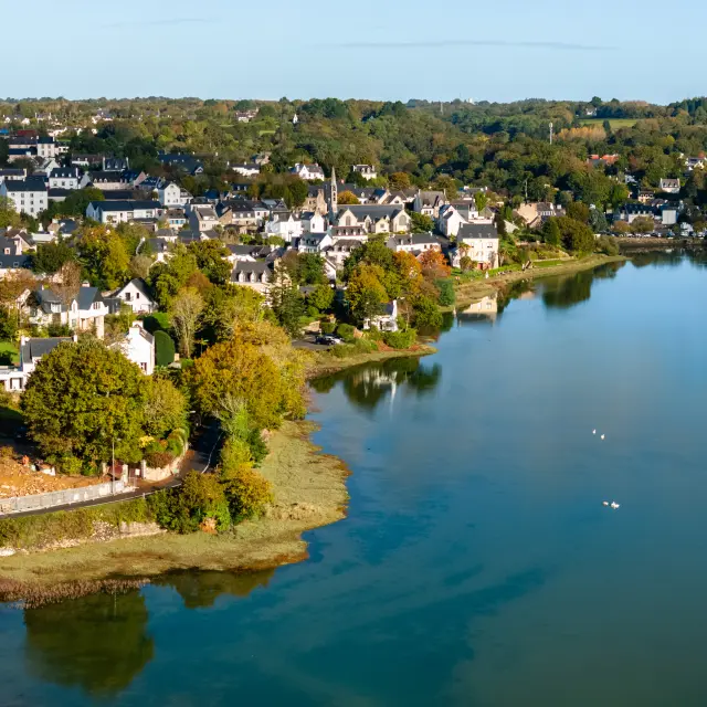 Une photo qui montre une vue aérienne d’un bourg au bord d’une grande étendue d’eau calme. Des maisons claires sont regroupées à gauche et au fond, entourées d’arbres verts et jaunes. La rive est bordée de végétation et d’un chemin/route, et l’eau occupe toute la partie droite avec quelques petits oiseaux à la surface.