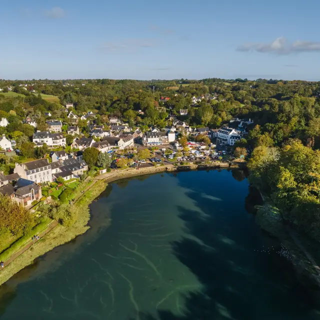 Une photo aérienne qui montre une anse bordée d’arbres, avec des maisons et bâtiments d’un village sur la rive gauche, beaucoup de verdure, et un ciel bleu avec quelques nuages.
