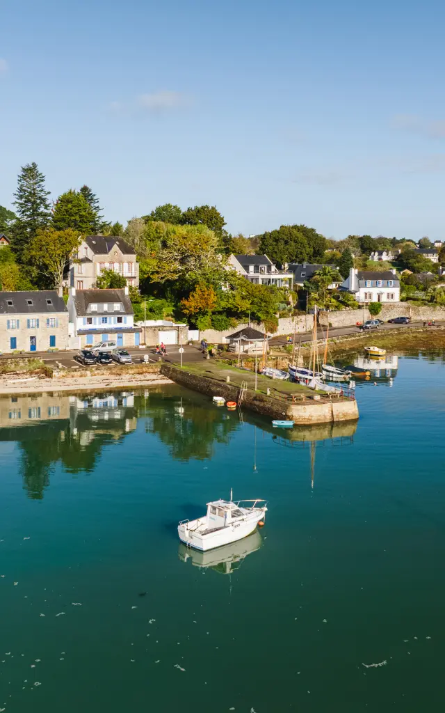 Une photo de port à gauche: eau calme bleu-vert avec plusieurs bateaux (petits bateaux et voiliers) au mouillage; au fond, une rive avec des maisons blanches/grises et des arbres verts et orangés; ciel bleu clair.