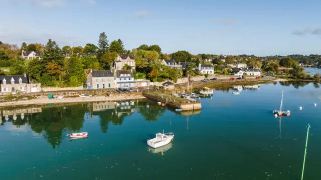 Une photo de port à gauche: eau calme bleu-vert avec plusieurs bateaux (petits bateaux et voiliers) au mouillage; au fond, une rive avec des maisons blanches/grises et des arbres verts et orangés; ciel bleu clair.