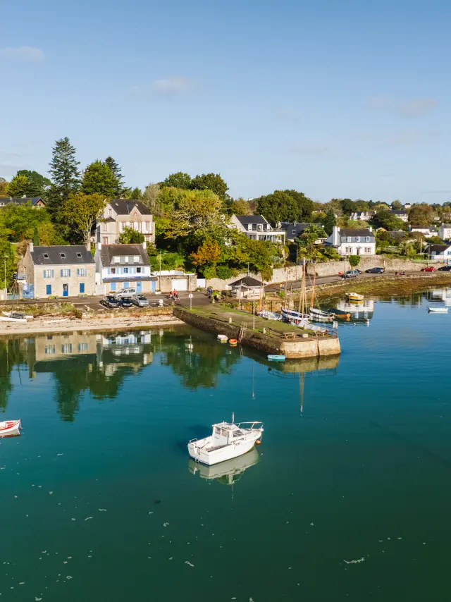 Une photo de port à gauche: eau calme bleu-vert avec plusieurs bateaux (petits bateaux et voiliers) au mouillage; au fond, une rive avec des maisons blanches/grises et des arbres verts et orangés; ciel bleu clair.