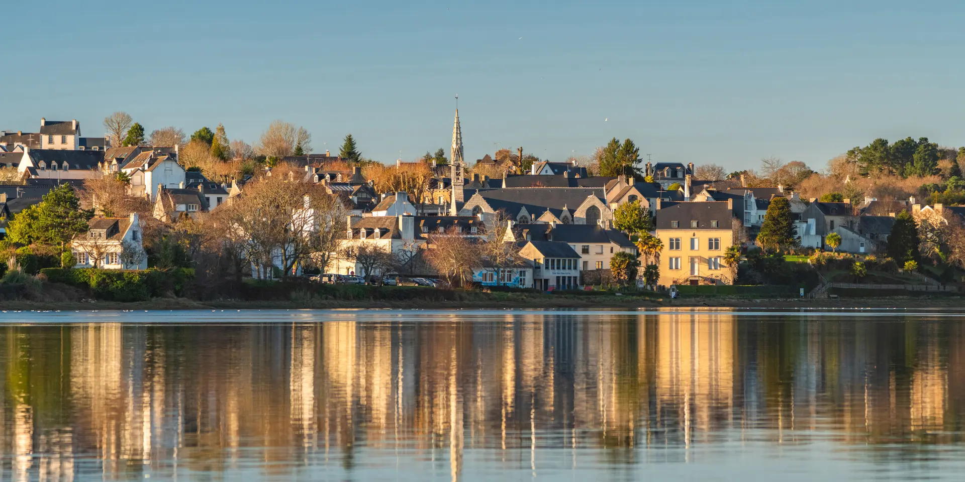Une photo qui montre un village au bord de l’eau sous un ciel bleu, avec des maisons, des arbres et un clocher d'église au centre. Les bâtiments se reflètent dans une eau calme au premier plan.