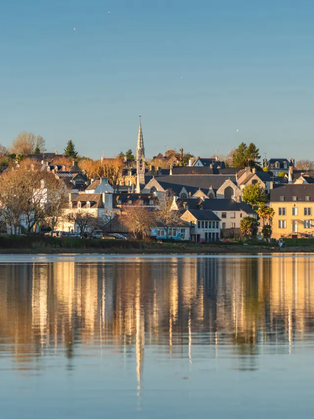 Une photo qui montre un village au bord de l’eau sous un ciel bleu, avec des maisons, des arbres et un clocher d'église au centre. Les bâtiments se reflètent dans une eau calme au premier plan.