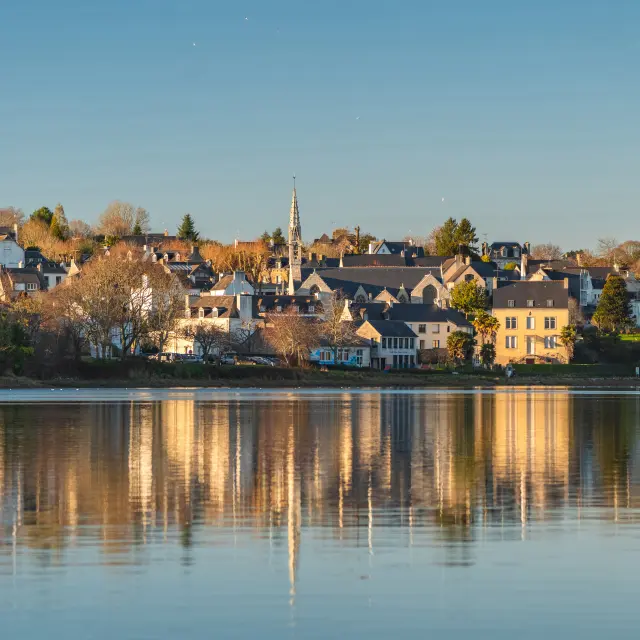 Une photo qui montre un village au bord de l’eau sous un ciel bleu, avec des maisons, des arbres et un clocher d'église au centre. Les bâtiments se reflètent dans une eau calme au premier plan.