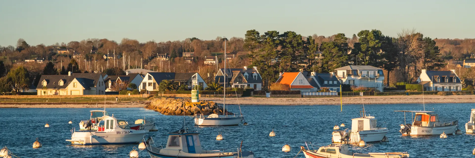 Une photo de bateaux au mouillage qui montre une étendue d’eau avec plusieurs petits bateaux (principalement des bateaux à moteur et un voilier) amarrés sur des bouées blanches, et au fond une rive avec des maisons et des arbres sous un ciel clair.