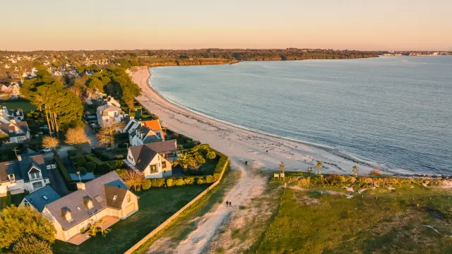 Une photo qui montre une plage courbe au bord de la mer, avec des maisons et des arbres près du rivage, lumière dorée de fin de journée, et une grande étendue d’eau calme à droite.