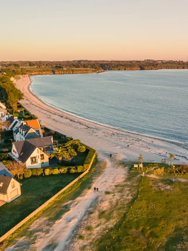 Une photo qui montre une plage courbe au bord de la mer, avec des maisons et des arbres près du rivage, lumière dorée de fin de journée, et une grande étendue d’eau calme à droite.