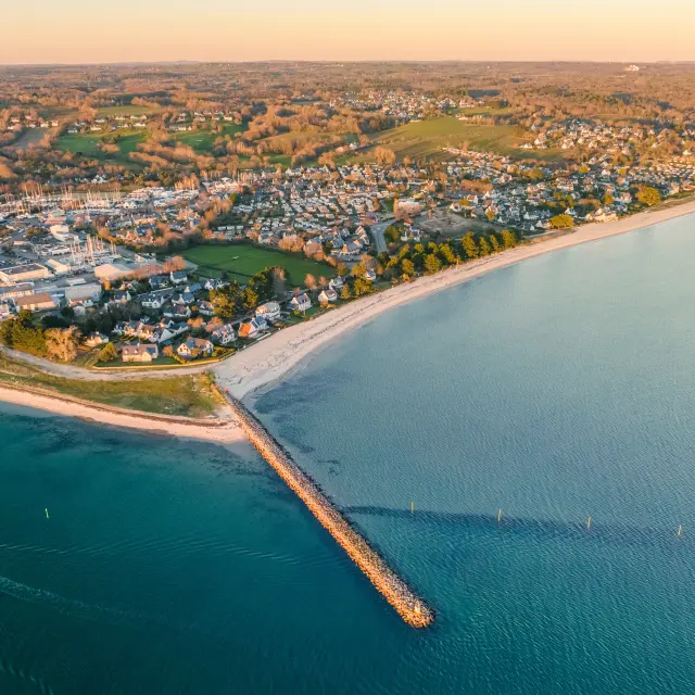 Une photo aérienne d’un littoral. La photo montre une plage en arc de cercle, une jetée s’avançant dans la mer, une mer calme et, à terre, un village avec des maisons et des zones vertes.