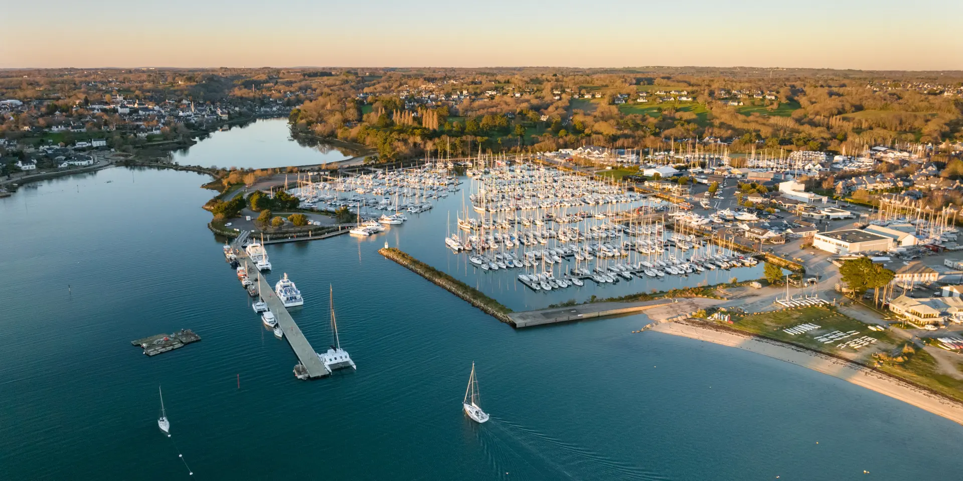 An aerial photograph showing a marina with numerous moored yachts, pontoons, a breakwater, a beach on the right, and a wide expanse of water in the foreground; in the background, dwellings and wooded areas under a clear sky.