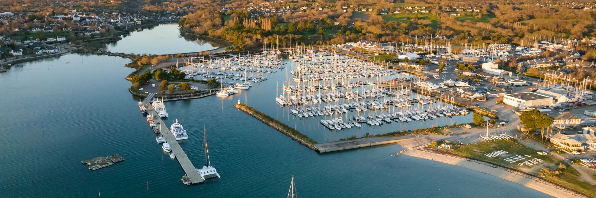 Une photo aérienne montrant un port de plaisance avec de nombreux voiliers amarrés, des pontons, une digue, une plage sur la droite, et une large étendue d’eau au premier plan; au fond, des habitations et des zones boisées sous un ciel clair.