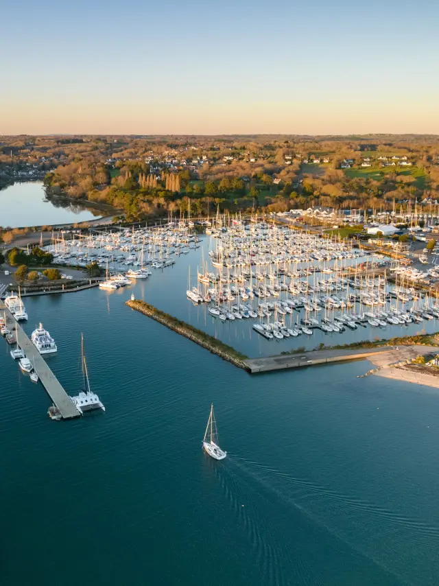 Une photo aérienne montrant un port de plaisance avec de nombreux voiliers amarrés, des pontons, une digue, une plage sur la droite, et une large étendue d’eau au premier plan; au fond, des habitations et des zones boisées sous un ciel clair.