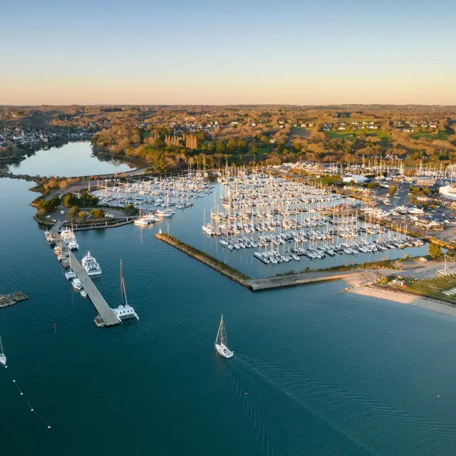 An aerial photograph showing a marina with numerous moored yachts, pontoons, a breakwater, a beach on the right, and a wide expanse of water in the foreground; in the background, dwellings and wooded areas under a clear sky.